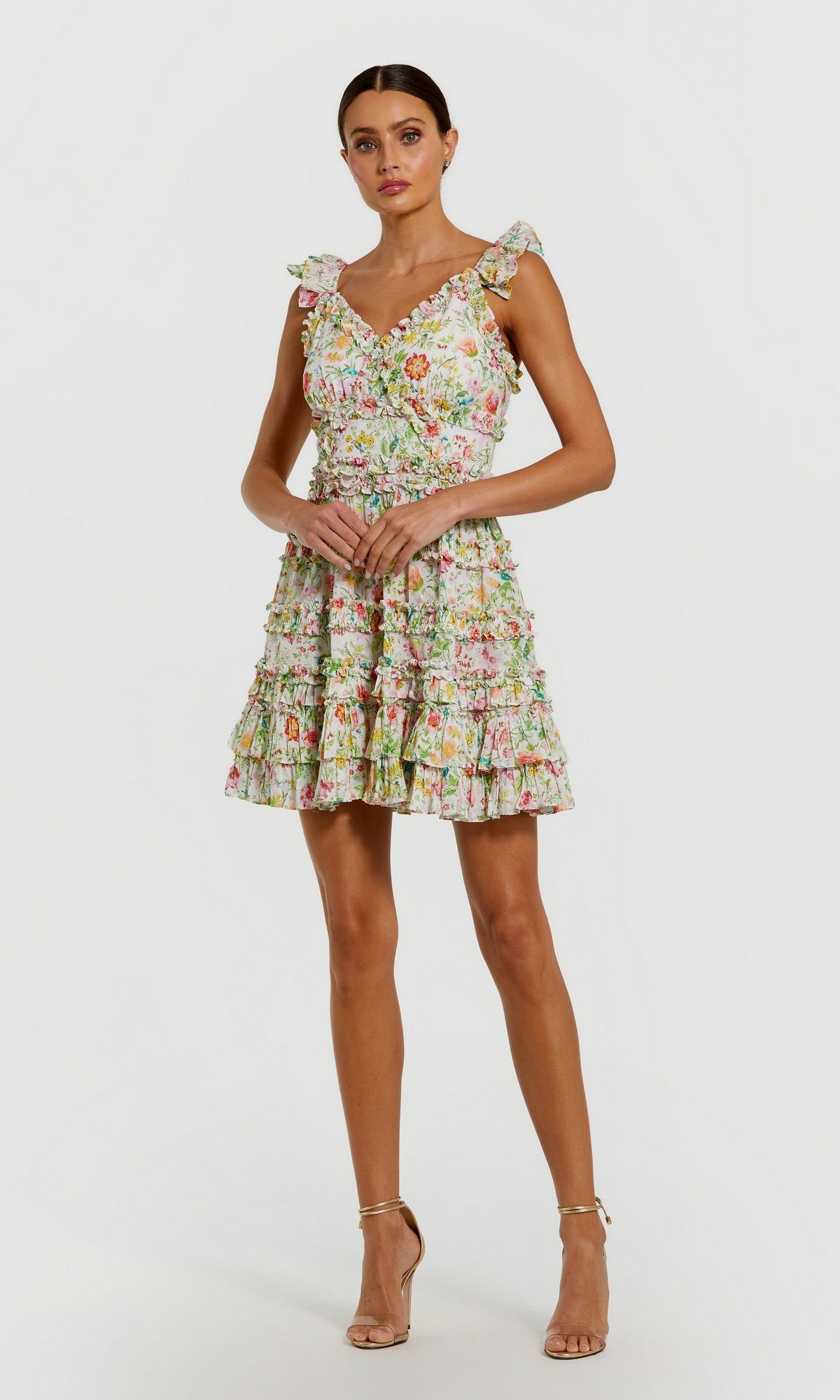 A woman wearing the Mac Duggal 8084 Short Party Dress in White Multi, featuring a sleeveless, ruffled, floral design, paired with strappy heels and standing against a plain white background.